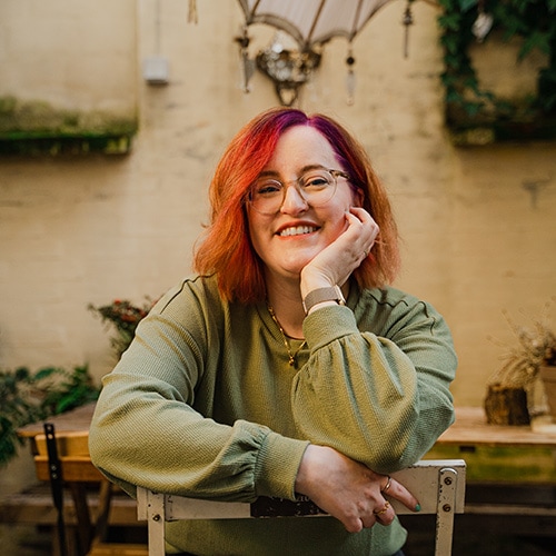 Birte Kahrs sitting on a backwards chair, leaning her right arm and her left elbow on the backrest, her chin in her left hand