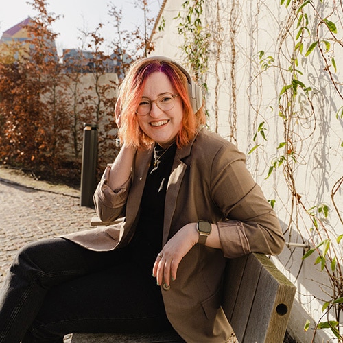 Birte Kahrs sitting at a bench outside, leaning her left elbow on the back rest and her right hand touching headphones on her head