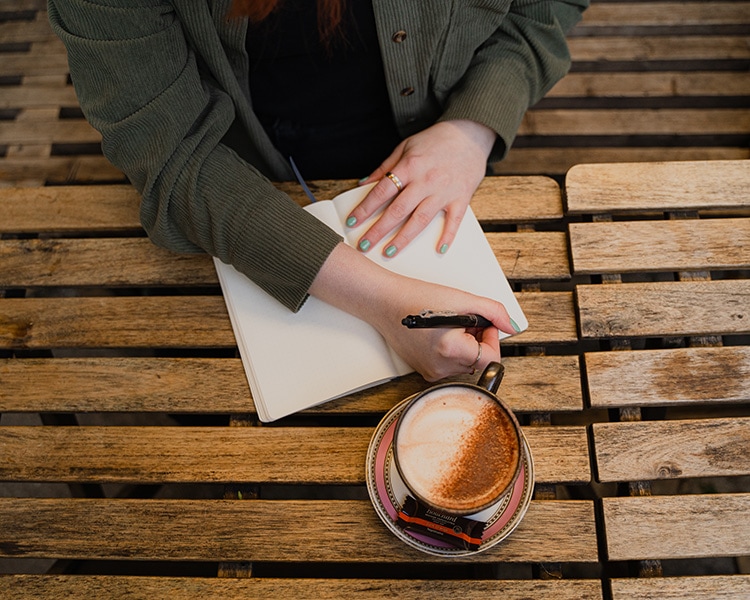 a top-down shot of an outdoor table that holds a cup of cappucchino and Birte is writing in a notebook with a pen