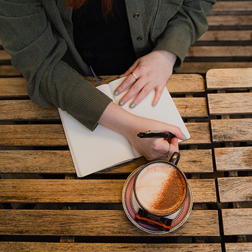 a top-down shot of an outdoor table that holds a cup of cappucchino and Birte is writing in a notebook with a pen