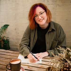 Birte Kahrs sitting at an outdoor table with a coffee, writing in a notebook with a pen