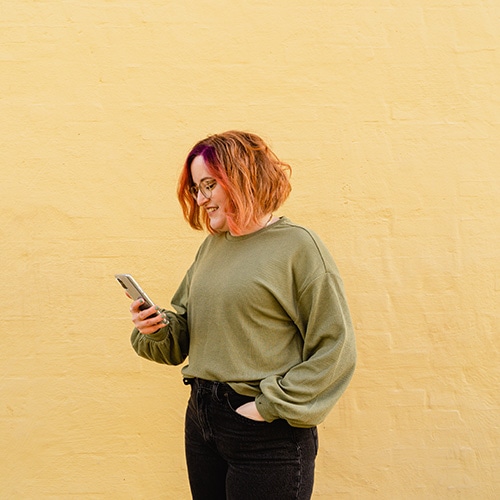Birte Kahrs standing in front of a yellow wall, looking at her phone in her right hand and her left hand in her pocket