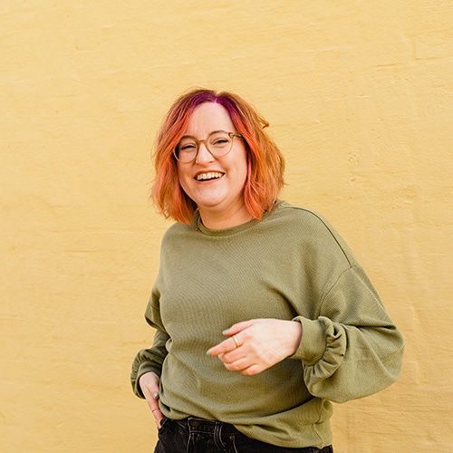 Birte Kahrs standing in front of a yellow wall, smiling and in motion with her hands